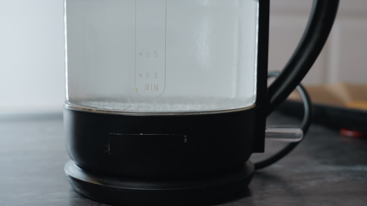 표면에 물 보일러를 놓고 있는 사람의 클로즈업, 물 수준과 항아리에 새겨진 글자에 초점을 맞추고, 가전제품과 쿠키 트레이가 있는 부의 일부를 볼 수 있습니다.