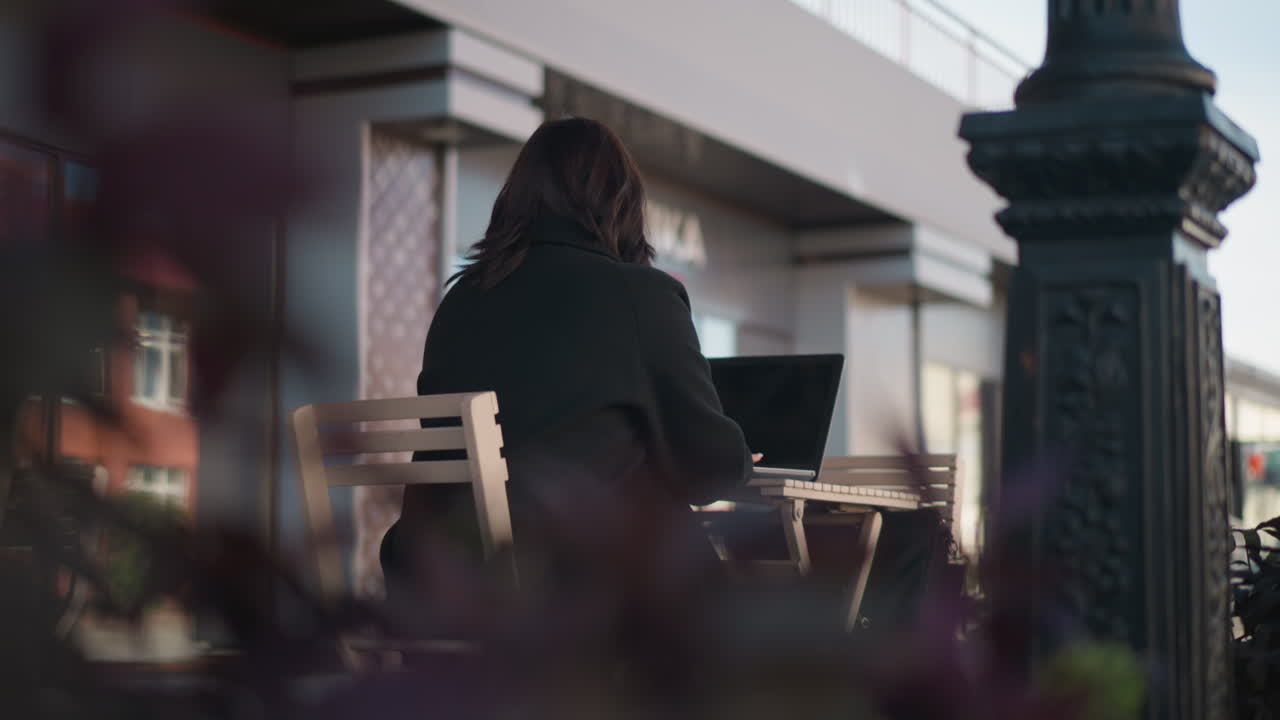Woman in black outfit typing on laptop while seated outdoors at restaurant table surrounded by wooden chairs and blurred plants in foreground, urban lifestyle scene