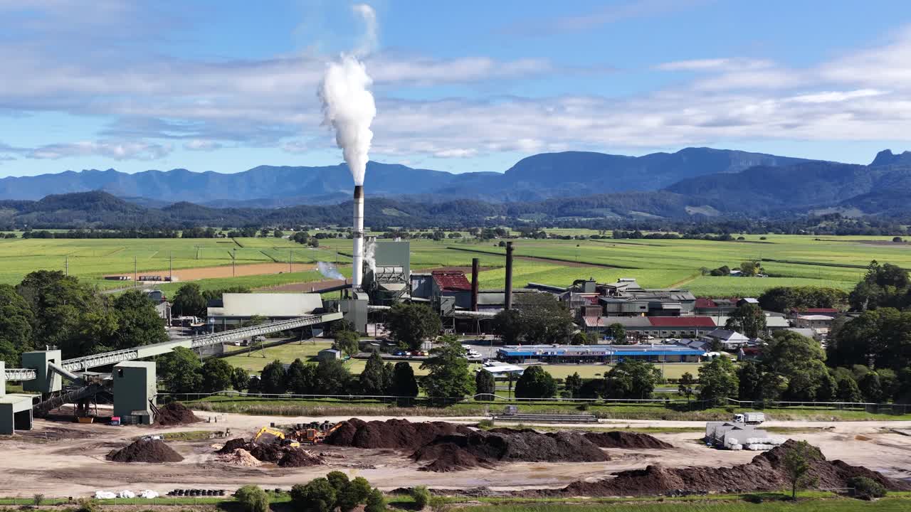 Aerial footage of a sugar mill with smoke stacks, surrounded by lush fields and mountains under clear skies