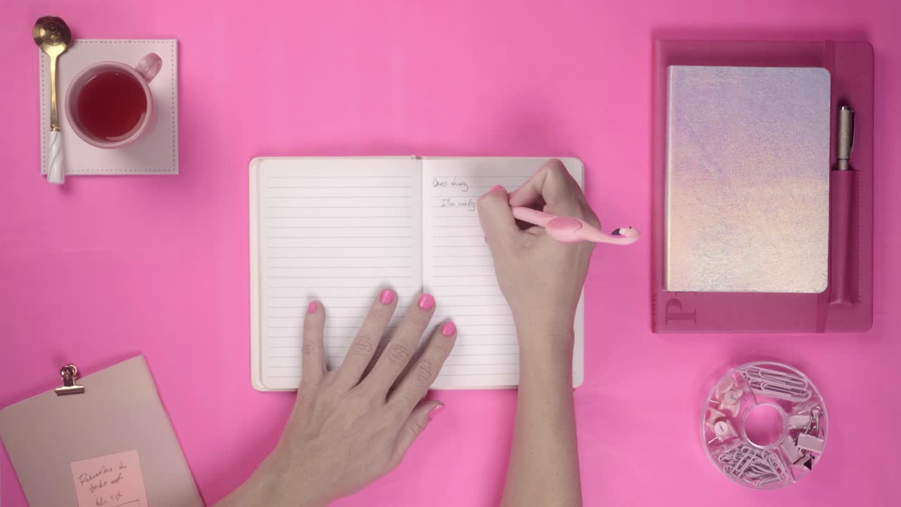 Top View of Hands Writing in a Notebook on a Pink Desk