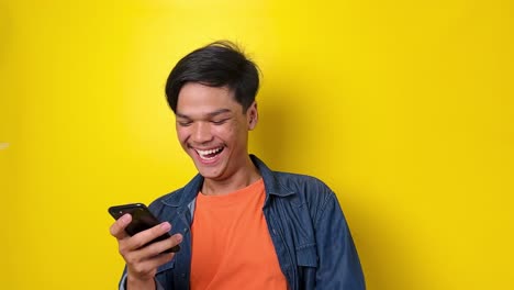 Young-Asian-man-in-casual-style-sees-something-very-funny-on-smartphone-and-laughs-out-loud,-isolated-on-yellow-background