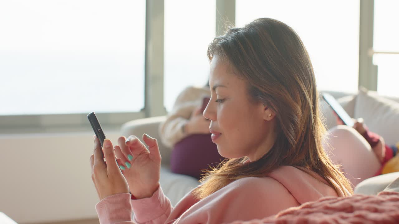 Mulher asiática de moletom rosa levantando o smartphone, ajustando a pegada e rolando as atualizações no sofá.
