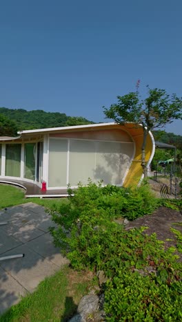vertical panning view of glam tree glamping resort with modern cabins, green lawn, hammock chair, and forested hills under clear blue sky