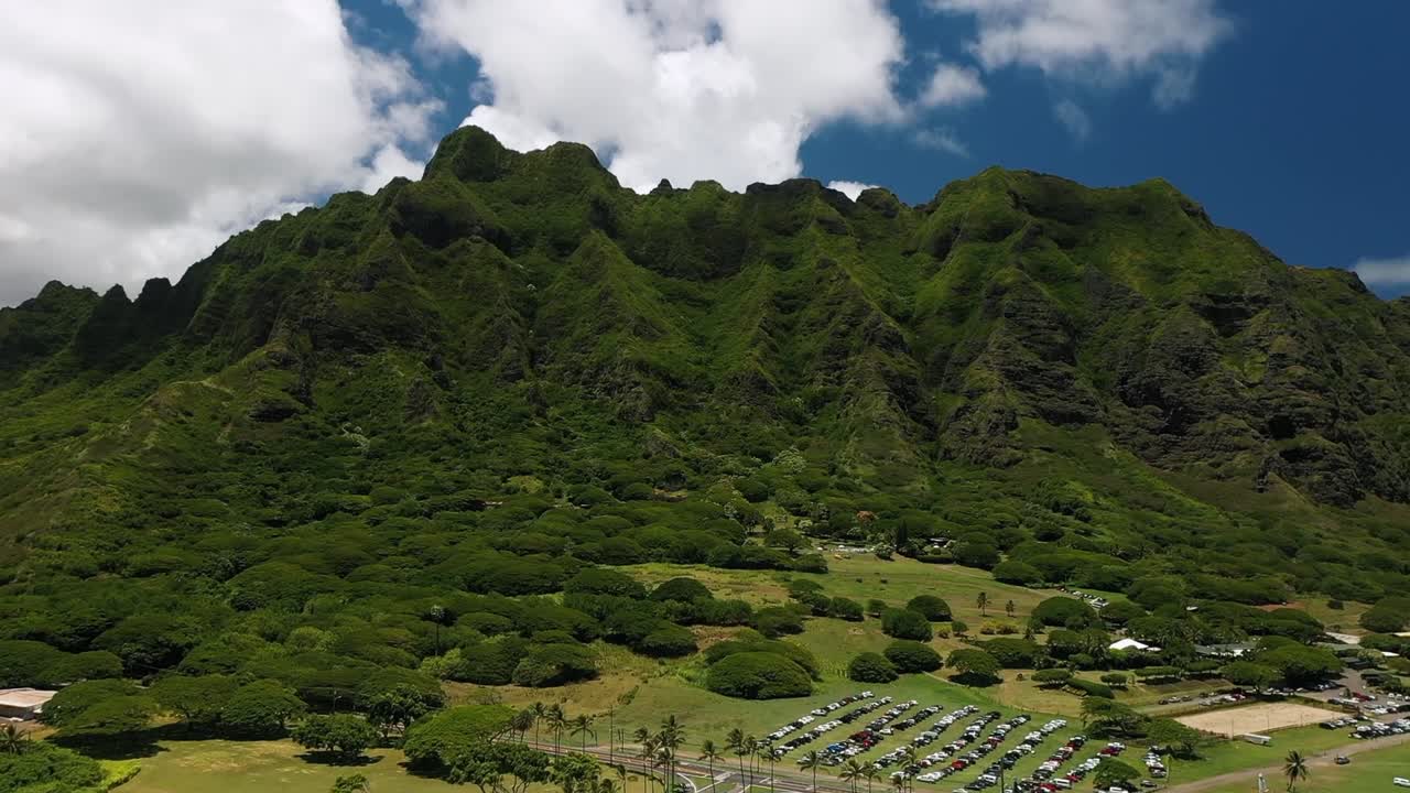 Aerial view showing lush green mountain and forest landscape of Lao Valley on Maui Island, Hawaii, with dense vegetation, open field, and parked cars under cloudy blue sky