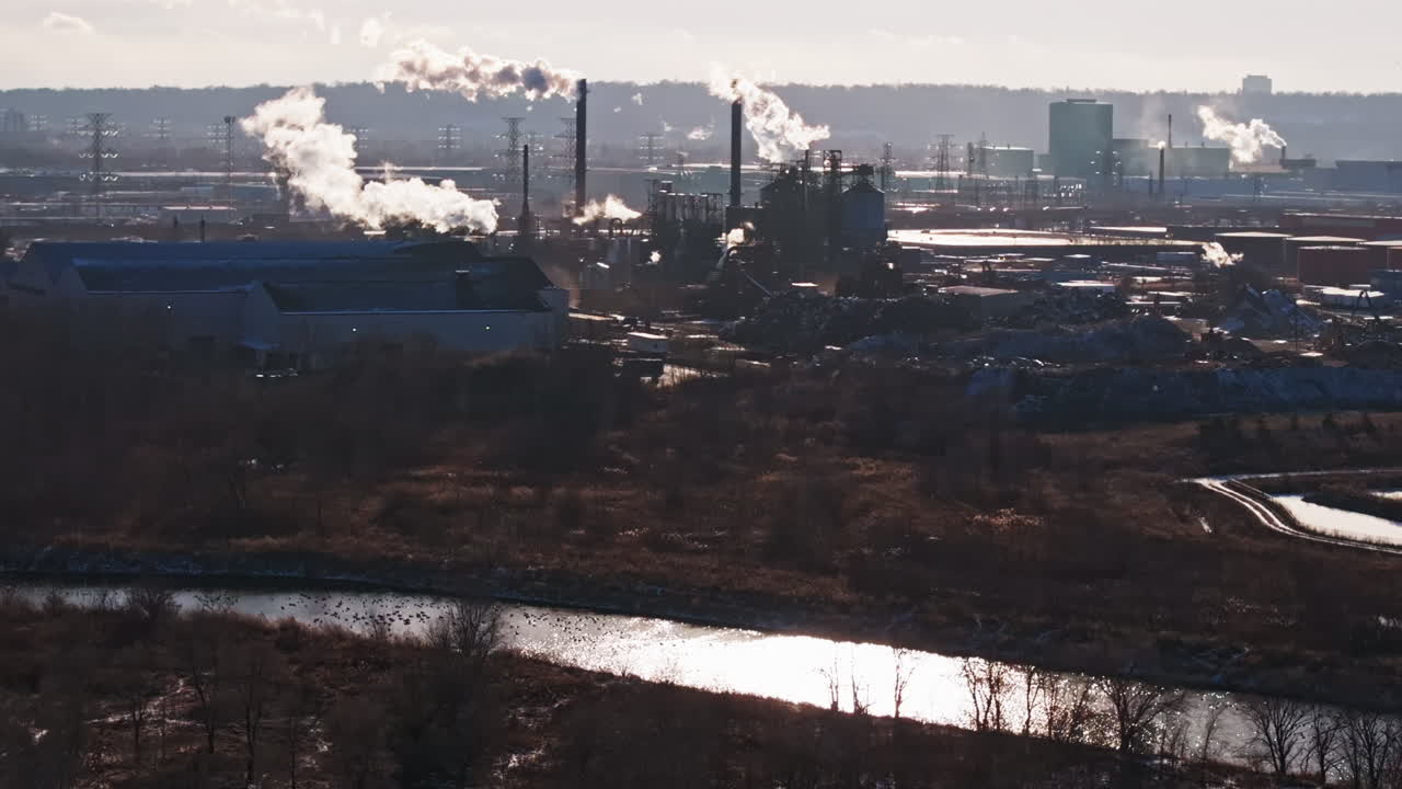 Industrial complex on riverbank with smokestacks emitting steam, backlit by sunlight, environmental impact concept