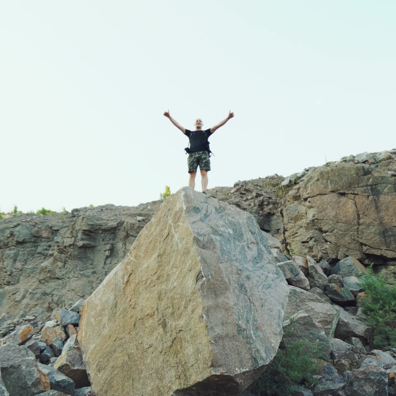 a brave traveler is standing on top of a huge rock with his hands raised on the background of a beautiful location in the summer