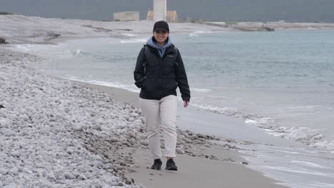 Mujer-Feliz-Caminando-Sola-En-Una-Playa-De-Ibiza,-Usando-Una-Chaqueta-Y-Una-Gorra---Toma-En-Cámara-Lenta