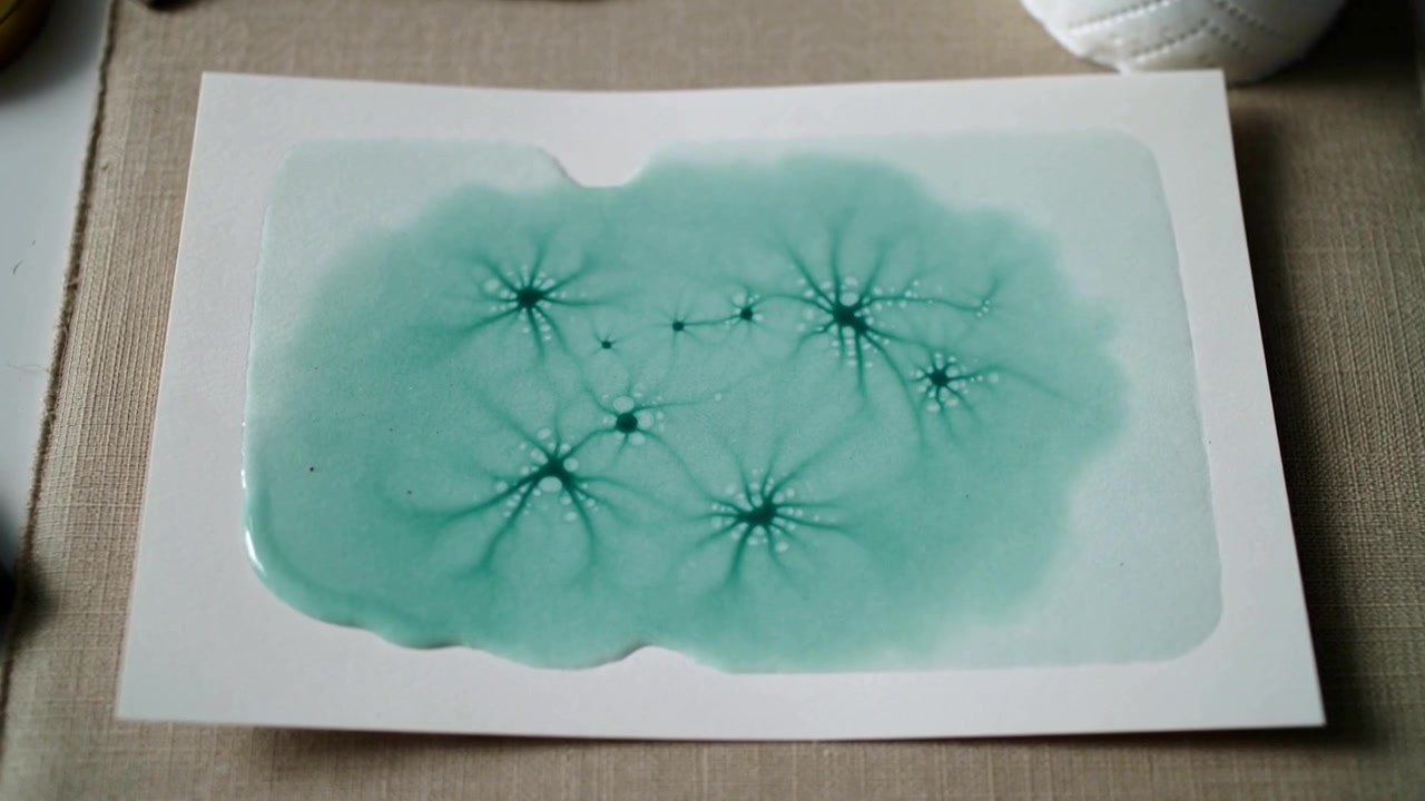 Verde acqua che si diffonde su un cartoncino posato su un tappetino intrecciato, con il centro che emette raggi a formare stelle, uno strumento di metallo e una palla trapuntata.