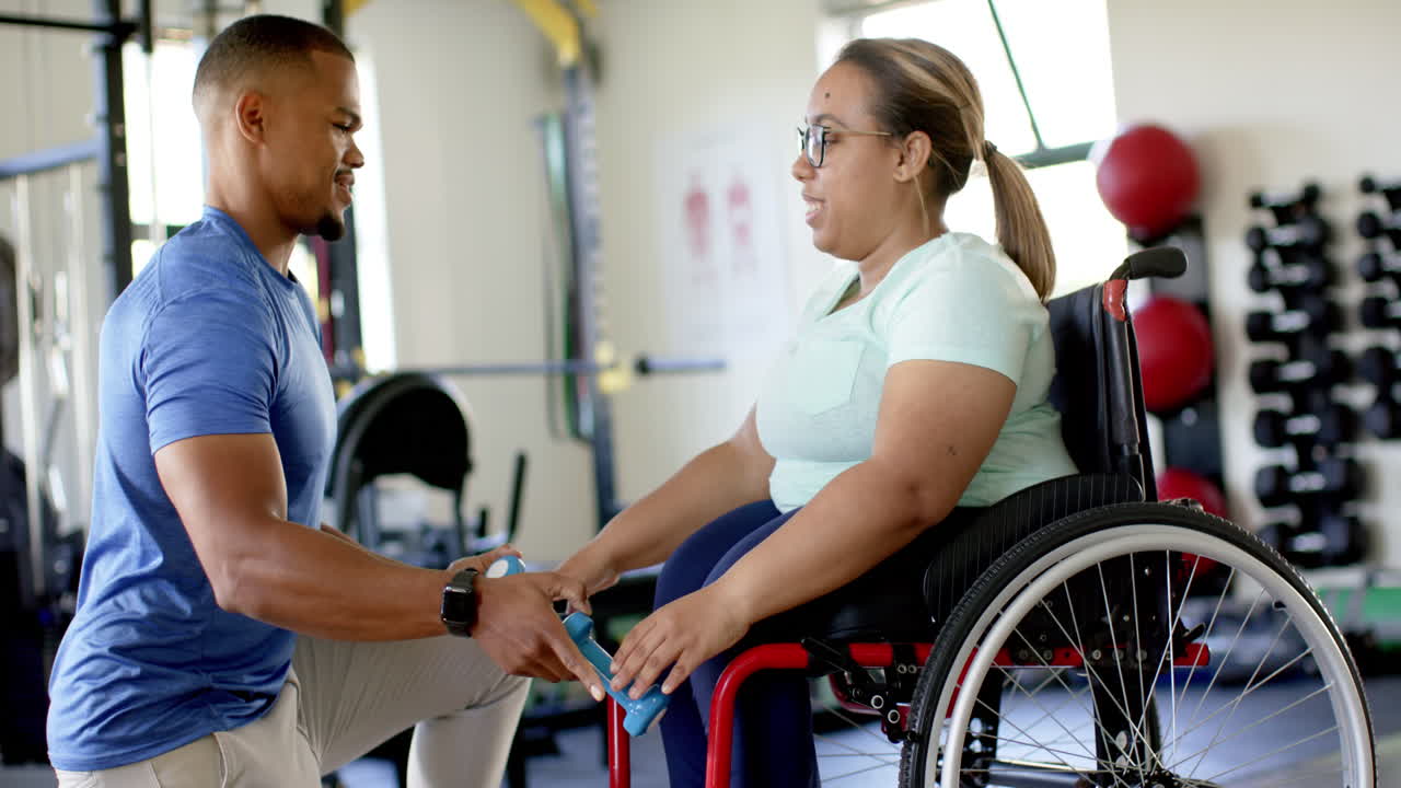 Physical therapist assisting woman with paraplegia in wheelchair with dumbbell exercises in gym