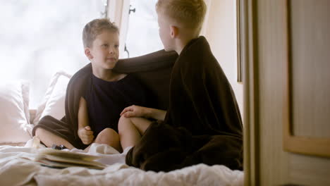 Two-little-brothers-with-blanket-on-their-shoulders-sitting-on-bed-in-a-campervan-and-talking-together