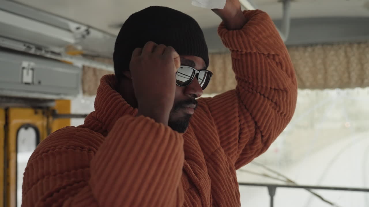 Uomo di colore alza le mani sull'autobus, indossa un berretto nero e un cappotto marrone oversize con occhiali da sole, espressione vivace e sicura, interno di un mezzo di trasporto pubblico, luce solare e riflesso sul finestrino, stile autentico