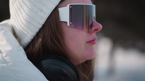 close up side view of young lady wearing headphones around neck and white knit beanie puts tinted goggles on face with manicured nails under sunlit clear winter sky breeze tousles hair