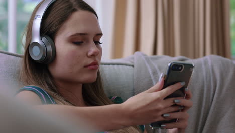 Fröhliches-Teenager-Mädchen,-Das-Mit-Kopfhörern-Musik-Hört-Und-Sich-Zu-Hause-Auf-Dem-Sofa-Entspannt-Und-Mit-Dem-Smartphone-SMS-Schreibt-Und-Dabei-Genießt,-Seinen-Lebensstil-In-Den-Sozialen-Medien-Zu-Teilen