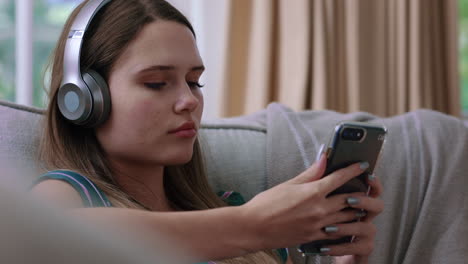 happy teenage girl listening to music wearing headphones relaxing on sofa at home texting with smartphone enjoying sharing lifestyle on social media