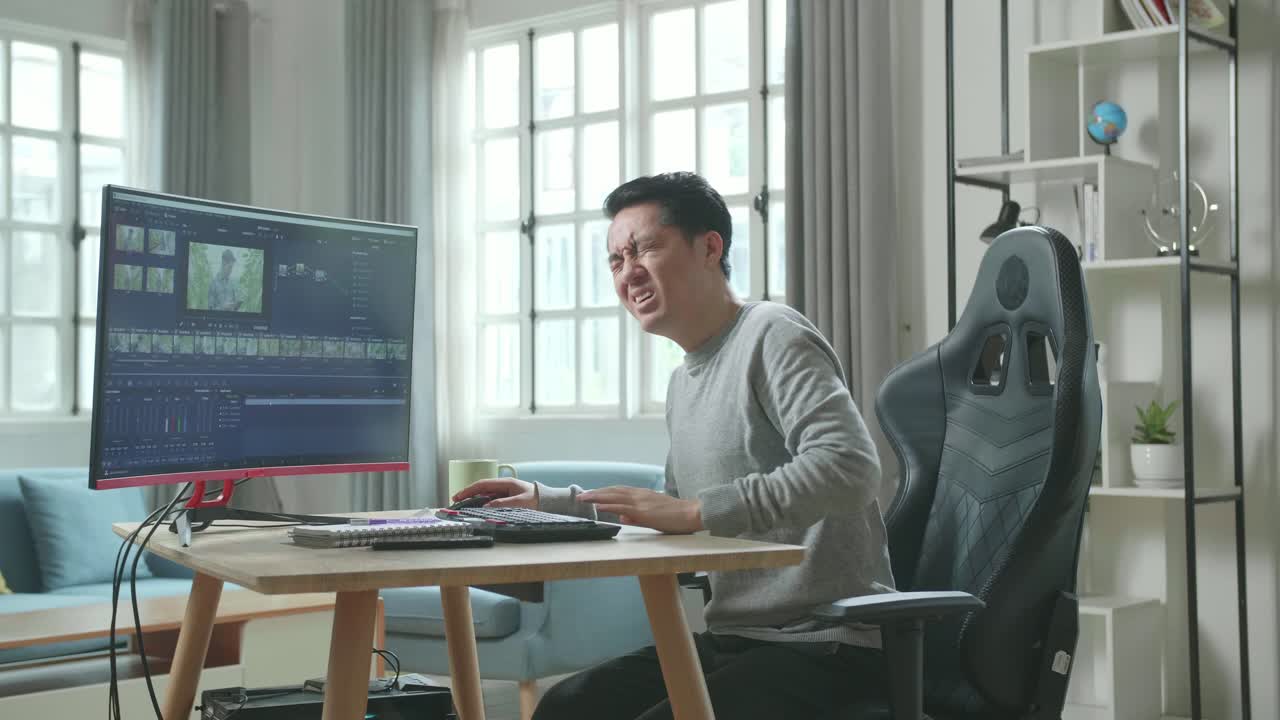 Asian Video Editor Man In Long Sleeved T-Shirt And Black Pants Having Backache While Using Desktop Computer For Editing Video At Home.