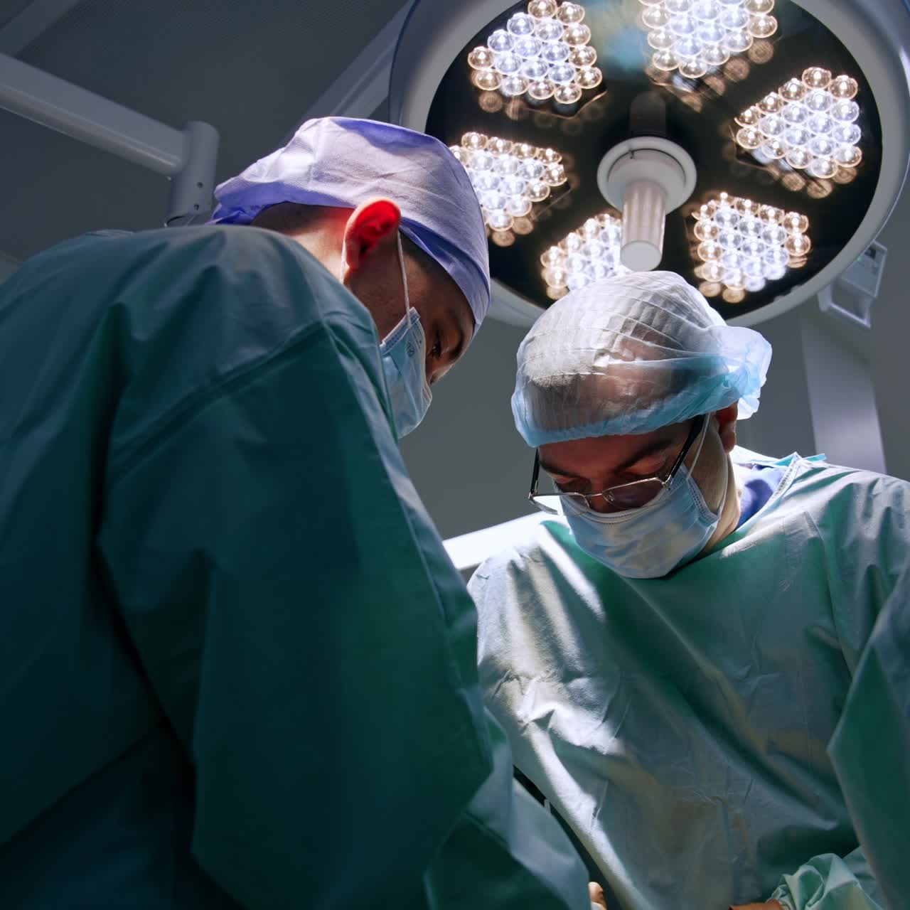 Medics in blue uniforms work in a team at operation. Low angle view. Round lamp on at the ceiling of the room at backdrop