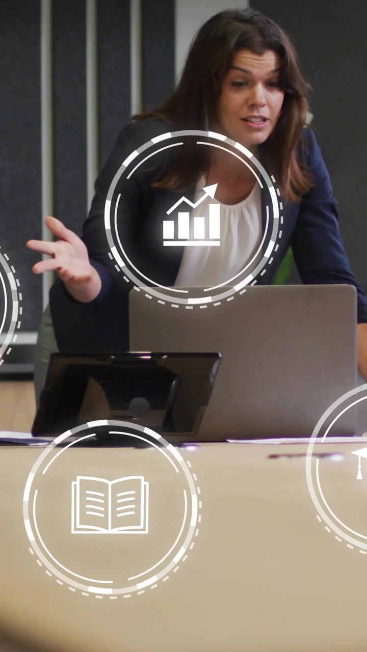 Donna di mezza età in blazer che conduce una riunione al tavolo dopo un segnale di un collega, indicando laptop, telefono e icone di grafici.
