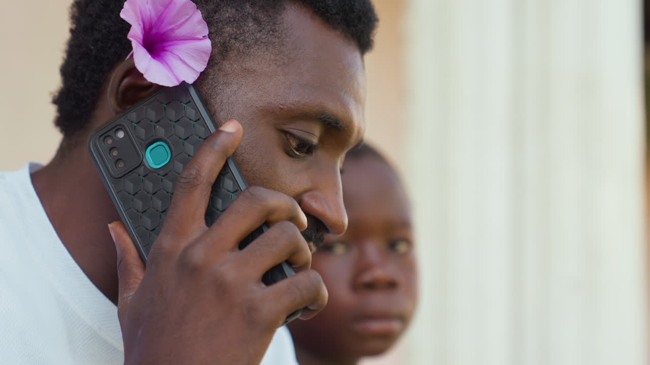 Black man on phone with purple flower, young boy watching behind, outdoor veranda portrait, thoughtful expression, warm afternoon light, textured phone case, close profile, community connection