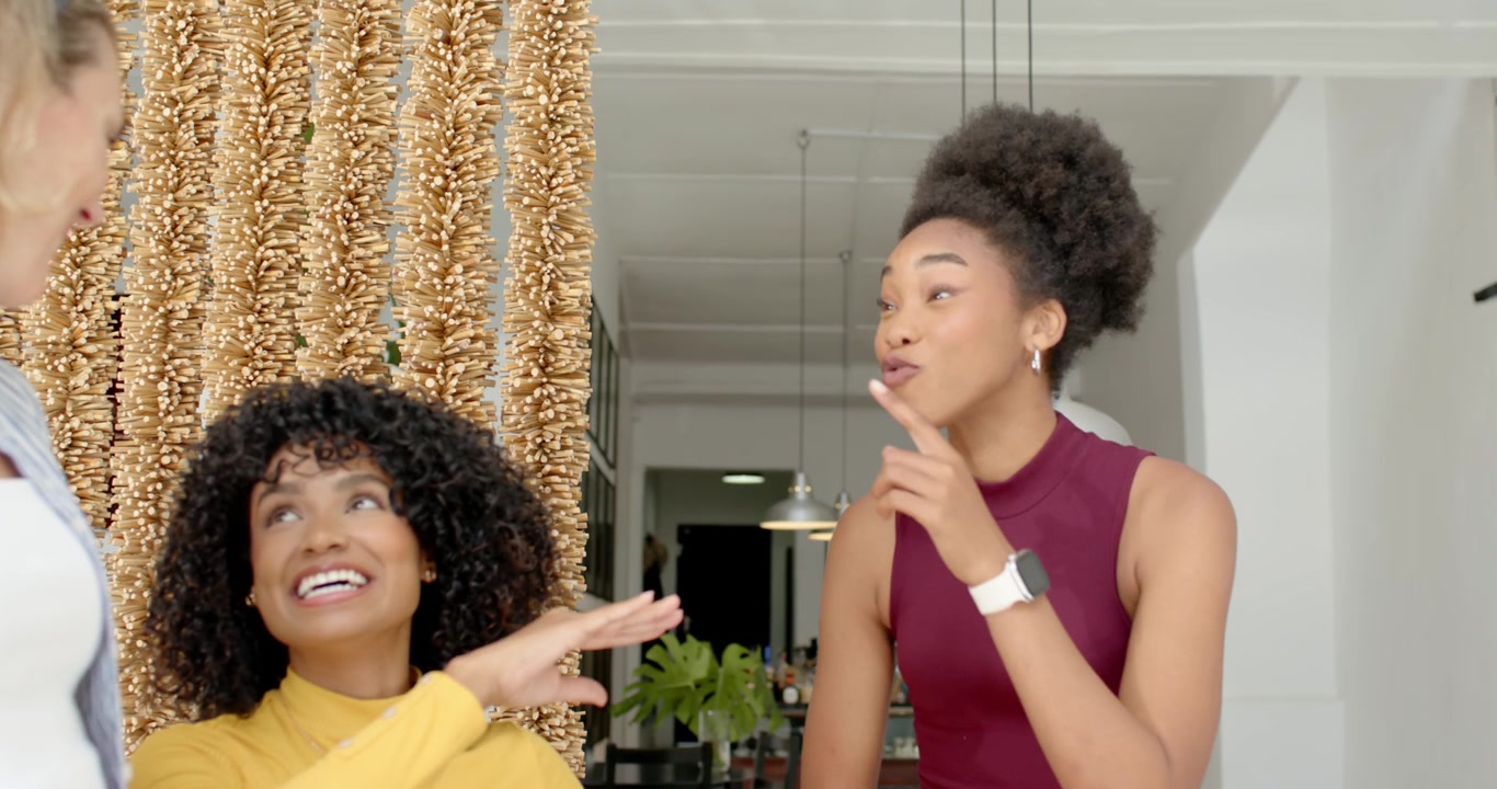 Diverse female friends in kitchen laughing pointing after seated woman raising hand near gold beads