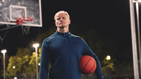 portrait of a confident basketball player holding ball and looking at camera while standing on an outdoor court at night