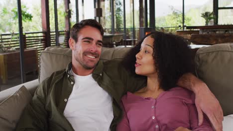 happy diverse couple sitting on couch in living room, watching tv, embracing and talking