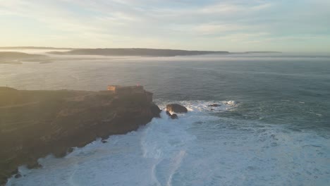 sunset view of a lighthouse on a cliff overlooking the ocean waves at farol de nazaré