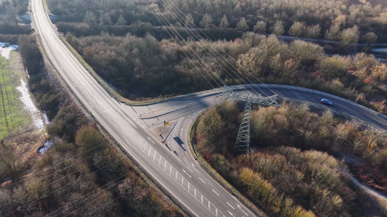 Vista aerea da drone di un incrocio stradale circondato da un paesaggio boschivo invernale, con linee elettriche ad alta tensione che attraversano la carreggiata. La scena mostra infrastrutture moderne che combinano i trasporti