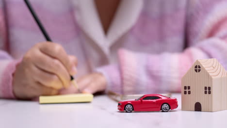 close-up of a hand holding a pencil and writing something on a yellow paper, behind a red toy car that signifies car loan financing