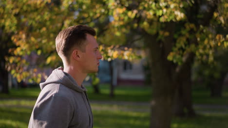 man walking through park with autumn foliage in background, wearing light gray hoodie and focused expression. soft sunlight filters through trees, highlighting greenery