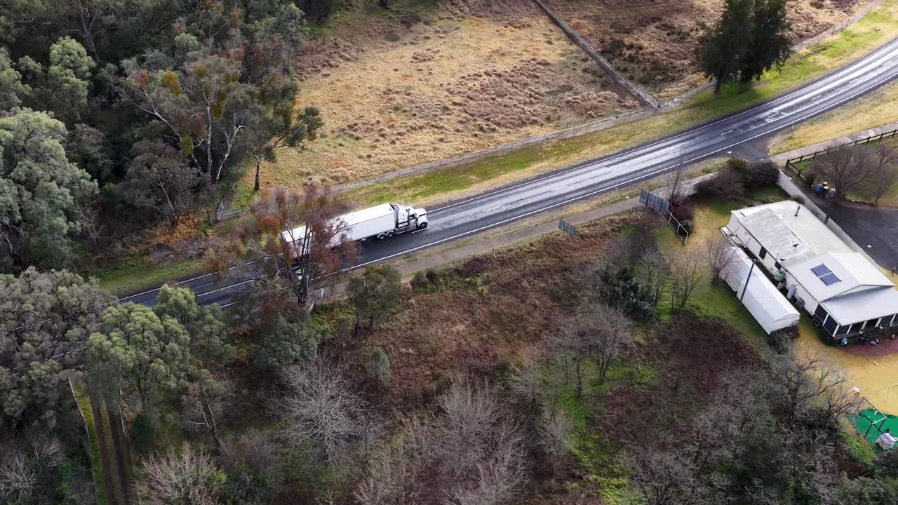 Two large trucks travel along a winding countryside road bordered by trees and rural homes, captured in smooth daylight aerial footage with steady camera movement