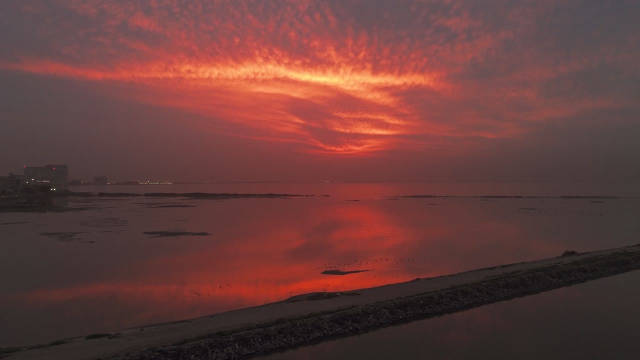 Weitwinkelansicht feuerroter Dämmerungswolken, die sich auf ruhigem Lagunenwasser spiegeln, mit dunkler Sandbank-Uferlinie und entfernten Gebäuden, die ein stimmungsvolles Küstenpanorama schaffen