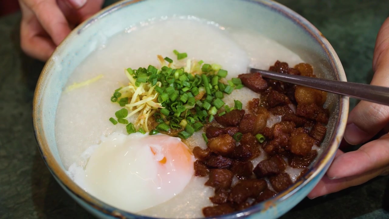 Close-up tangan memutar semangkuk bubur nasi gurih dengan telur dan daging babi.