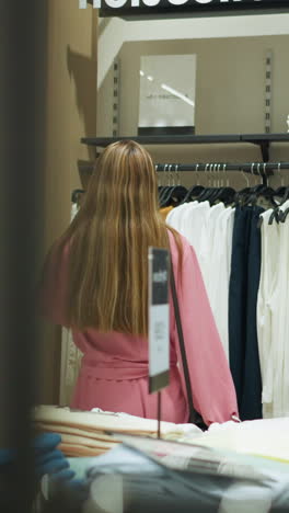 vista trasera de una dama blanca con una camiseta rosa y pantalones cortos, navegando por la ropa bien colgada en una tienda, otros artículos de ropa están plegados y dispuestos en la mesa, en la tienda bien organizada