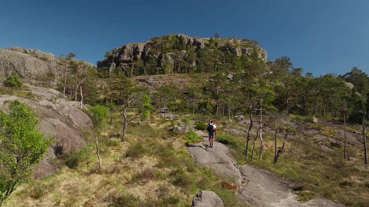 Drone sobe atrás de um casal de caminhantes em terreno rochoso revelando dramáticas montanhas de granito sob um céu azul claro ao longo da cênica trilha Hatten na Noruega. Paisagem lunar