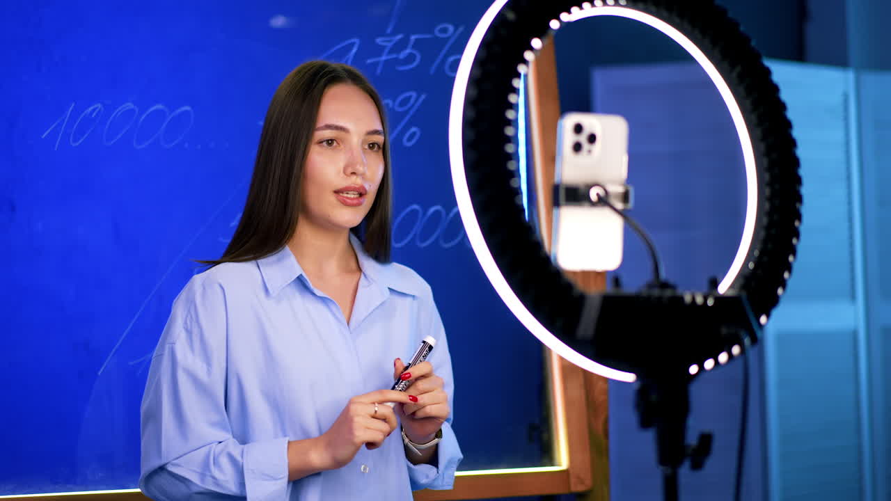 Good-looking Caucasian brunette standing at the glass board talks to camera. Blogger taking footage on the phone with ring light.