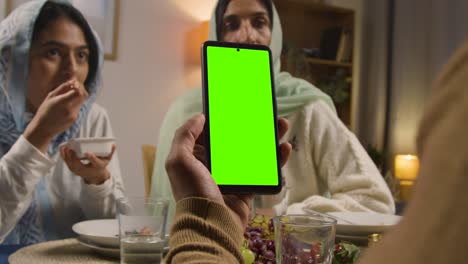 man looking at green screen mobile phone sitting around muslim family table at home eating iftar meal breaking daily fast during ramadan 1