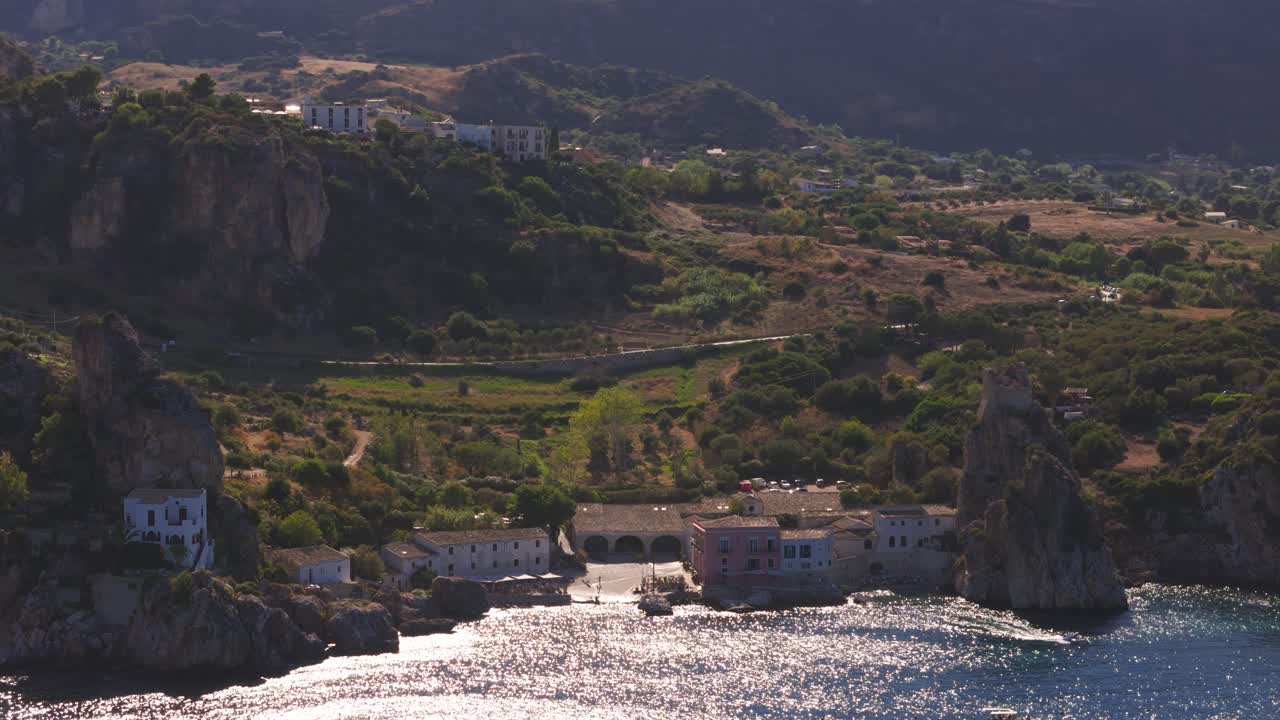 Aerial drone shot panning left above the calm blue waters at Tonnara di Scopello, Sicily, Italy, gliding over numerous boats as the ocean glistens under the warm golden sunlight during Italian summer