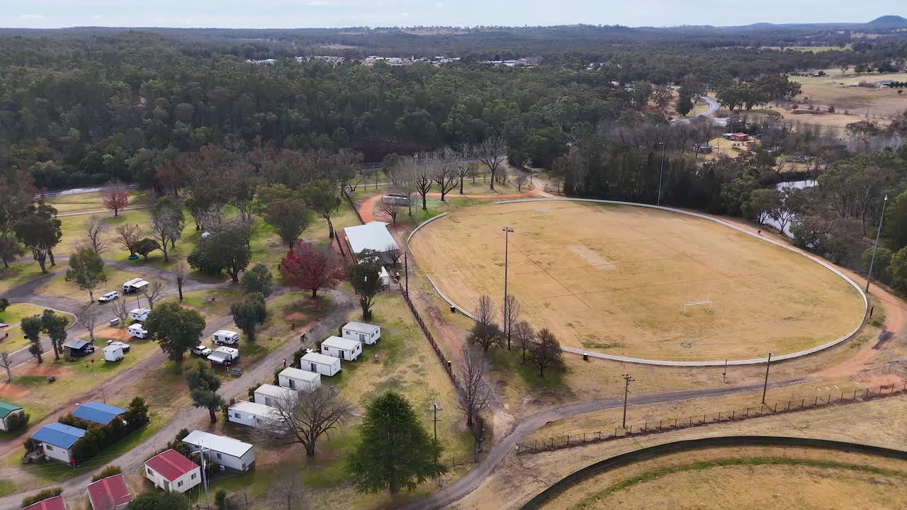 Drone camera glides above a dry sports field and nearby residential area in a rural Australian neighborhood, under overcast daylight with steady movement