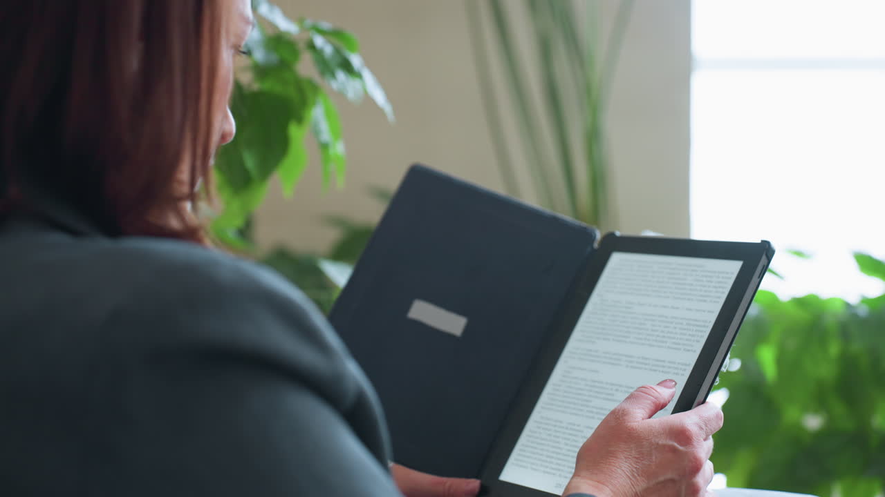 Mature woman using finger to scroll or turn page on digital e-reader while enjoying quiet reading moment indoors surrounded by green houseplants and soft natural light in peaceful cozy home setting