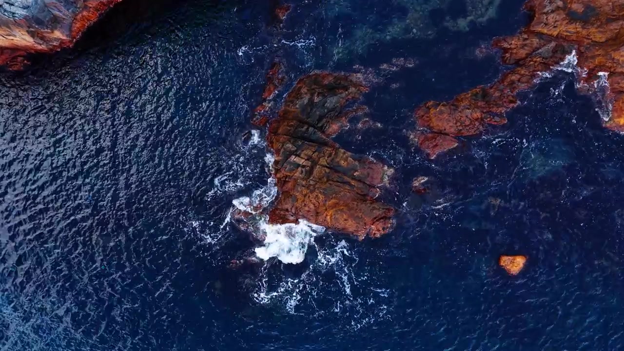 Waves breaking over rusty coastal rocks from above. White foam splashing against eroded red and orange stone in deep navy water, drone view.