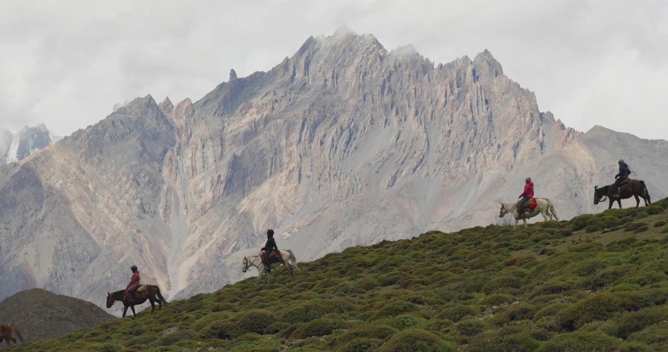 Aerial shot tracks across the green hills of Mustang, Nepal, moving in the opposite direction of horseback riders as they travel beneath a towering, rugged Himalayan peak