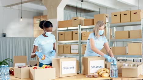 african american and caucasian women volunteers in facial masks packing donation boxes with food and water in charity warehouse