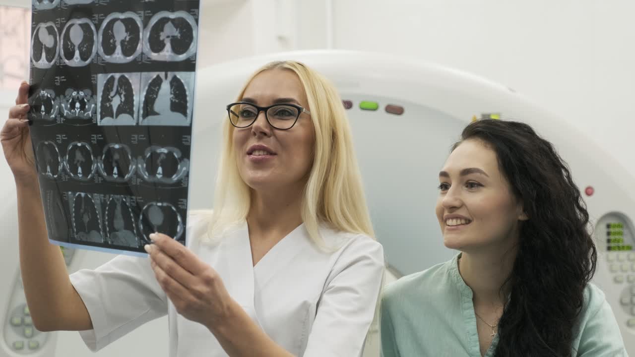 Woman doctor radiologist explains good results of CT scanning for young female patient, showing the snapshot with images, observing and analyzing CT scan in modern clinic beside modern CT scanner