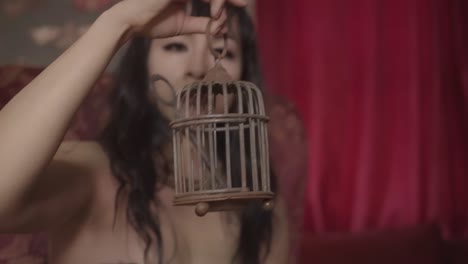 alternative girl with black hair sitting in a big red armchair in a gothic room holding a small bird cage with a key