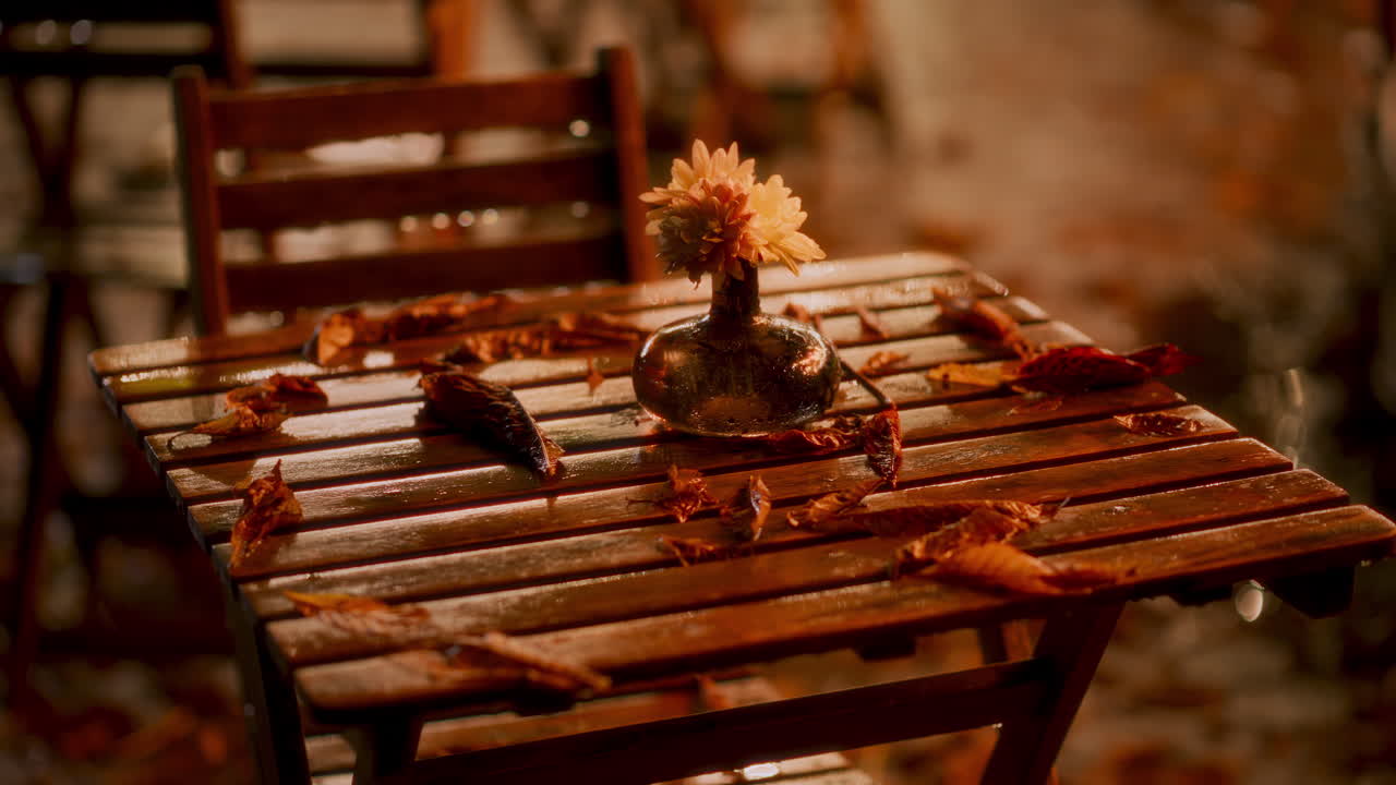 Raindrops gently hit a wooden table surrounded by colorful autumn leaves. A simple flower vase adds charm to the cozy outdoor setting on a rainy day