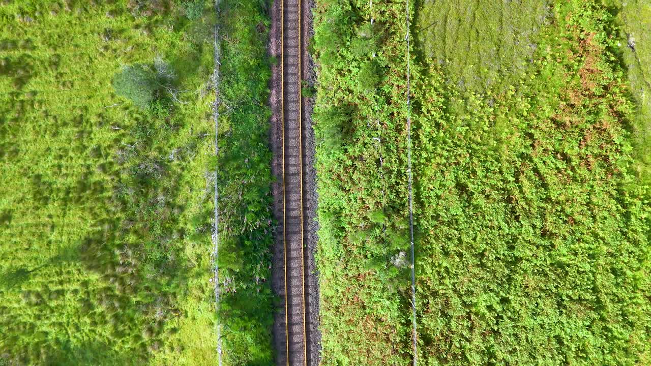 Drone footage moves steadily above single railway track through lush green Highlands landscape, midday sunlight casting soft shadows, wide overhead perspective