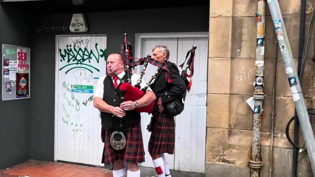 A bagpiper in traditional attire plays music beside a graffiti-covered door and stone wall.