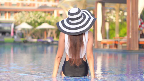 a young, fit woman in a bathing suit and huge black and white sun hat sits in the shallow end of the pool, enjoying the resort life