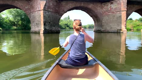 girl paddles canoe on river wye through stone bridge arch in the sun