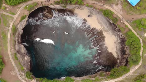 aerial cinematic view of broken beach circular rock formation with ocean waves crashing into the centre on edge of indonesian island during warm overcast day with blue water-1